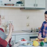 Young mother talking photo on smartphone camera of her cute daughter cooking cutting vegetables in the kitchen at home. Family, cook, and people concept